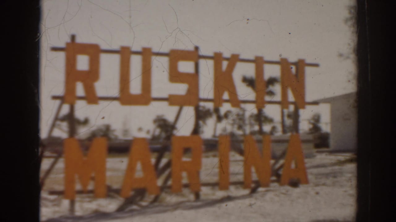 1963 Florida home-movie frame showing a large orange Ruskin Marina sign near pavement and sparse trees.