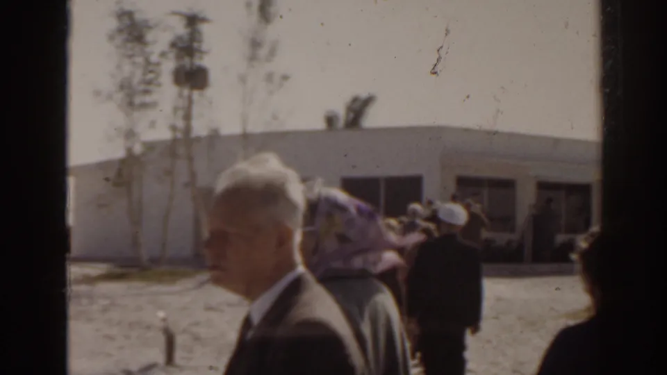 1963 Florida home-movie frame of an older suited man and other adults outside a low marina building.