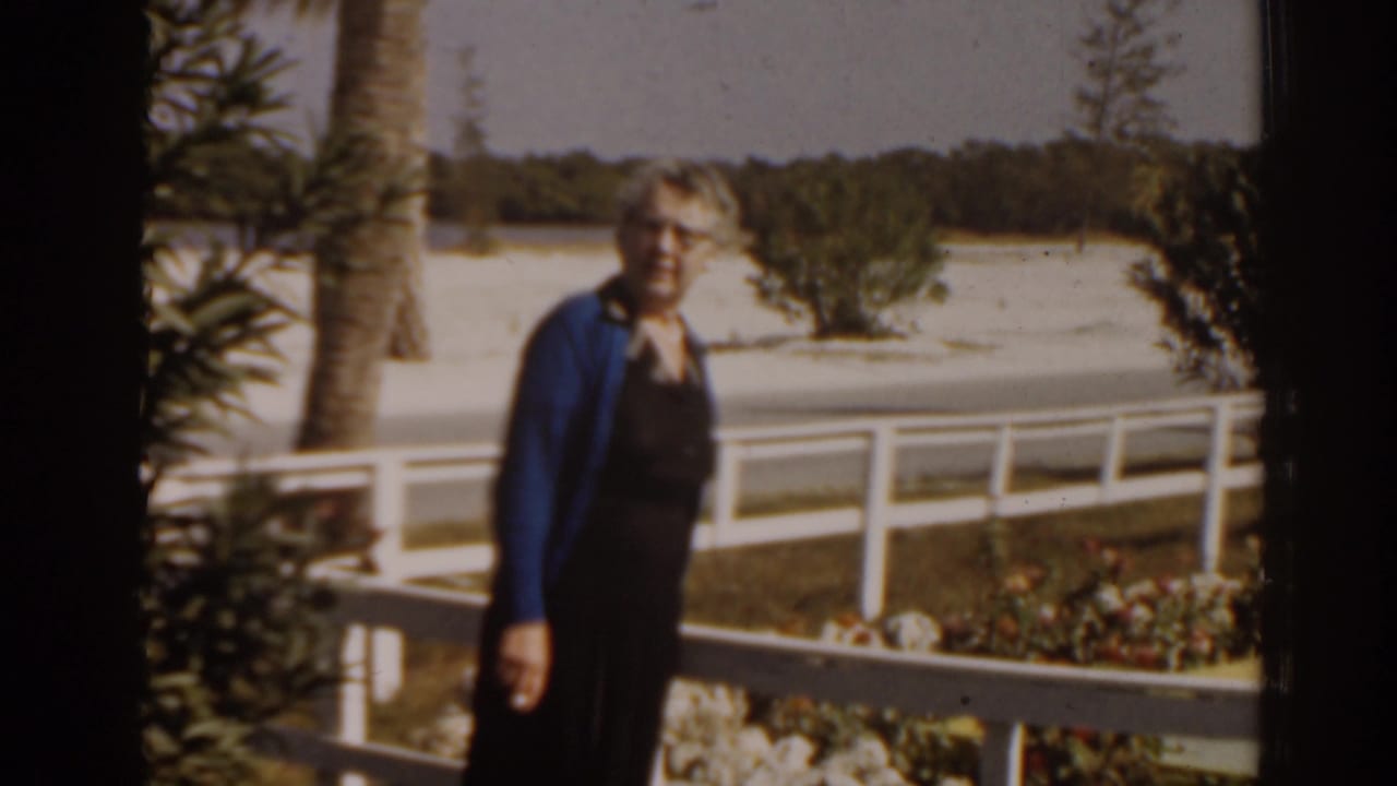 Elderly woman in a blue cardigan stands by a white railing outdoors.