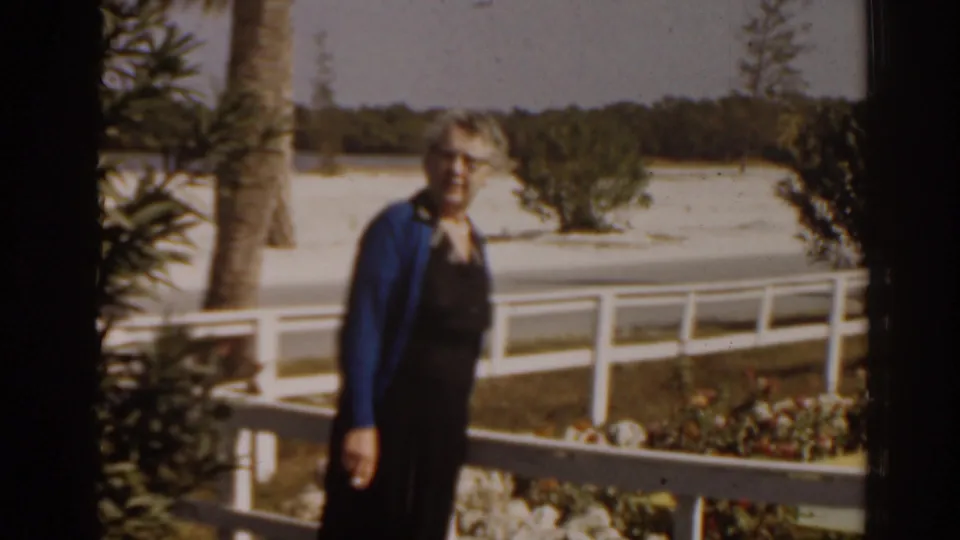 Elderly woman in a blue cardigan and dark dress stands near a white railing in a 1963 Florida home-movie frame.