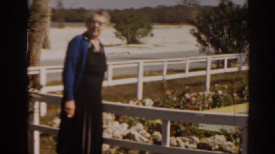 1963 Florida home-movie frame of a woman in a blue cardigan standing beside a railing, flower bed, palm trunk, and sandy background.
