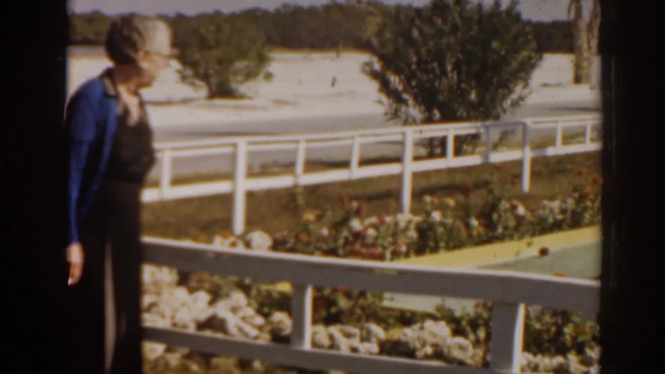 Woman in a blue cardigan turns slightly beside a white railing in the 1963 Florida home-movie scene.