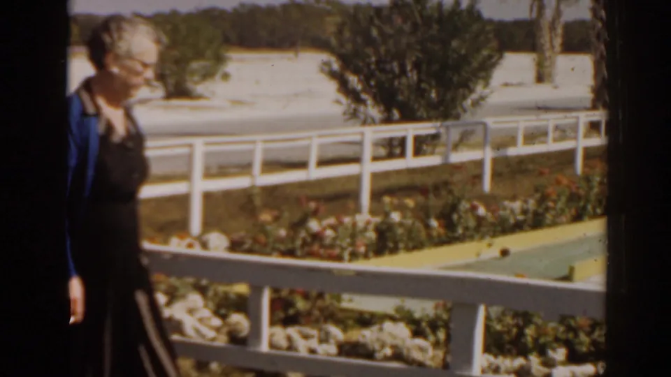 In 1963 Florida home-movie footage, an older woman stands near a window as pastel mini-golf greens and white railings appear outside.
