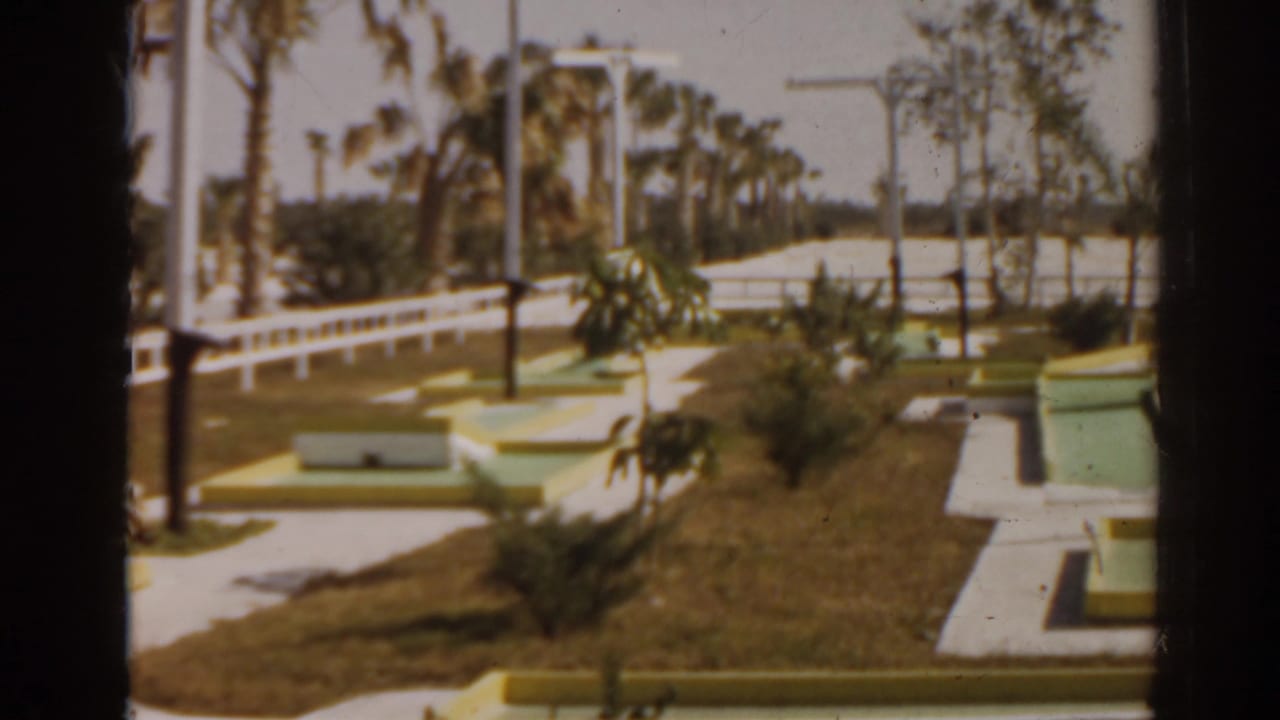 Pastel mini-golf course with white railings, palms, grass, and bright outdoor light.
