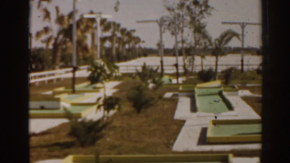 The 1963 Florida home-movie frame shows a landscaped mini-golf layout with pastel obstacles, white paths, palms, and roadside railings.