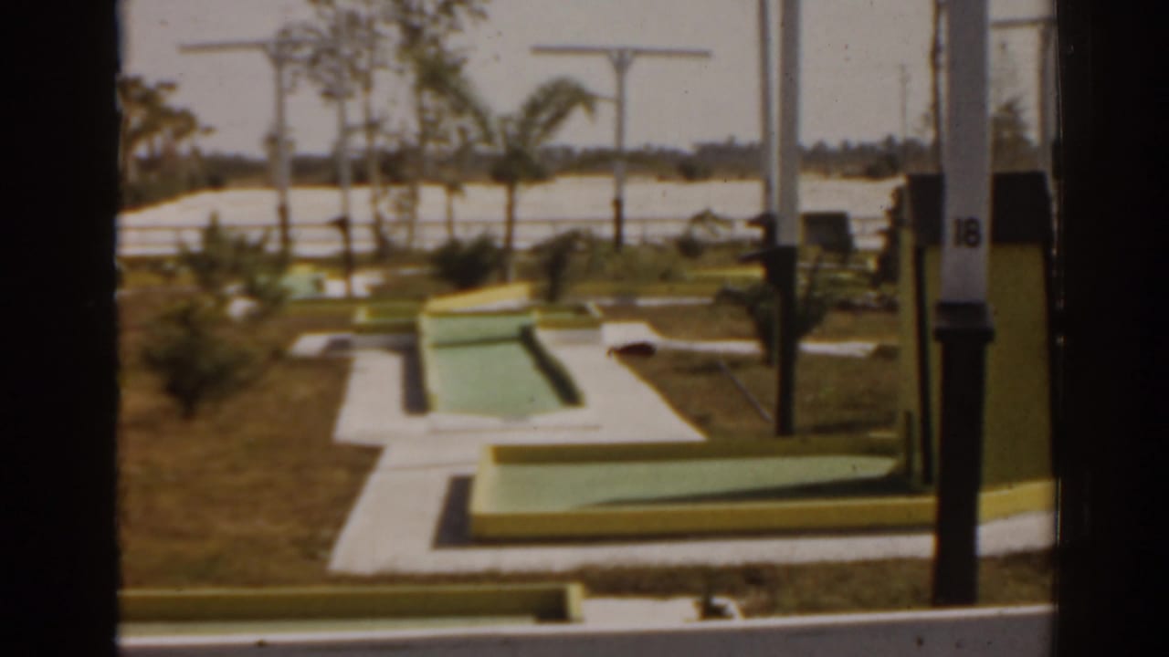 Miniature golf course with green putting surfaces, grass, palms, and a small road or waterway beyond.