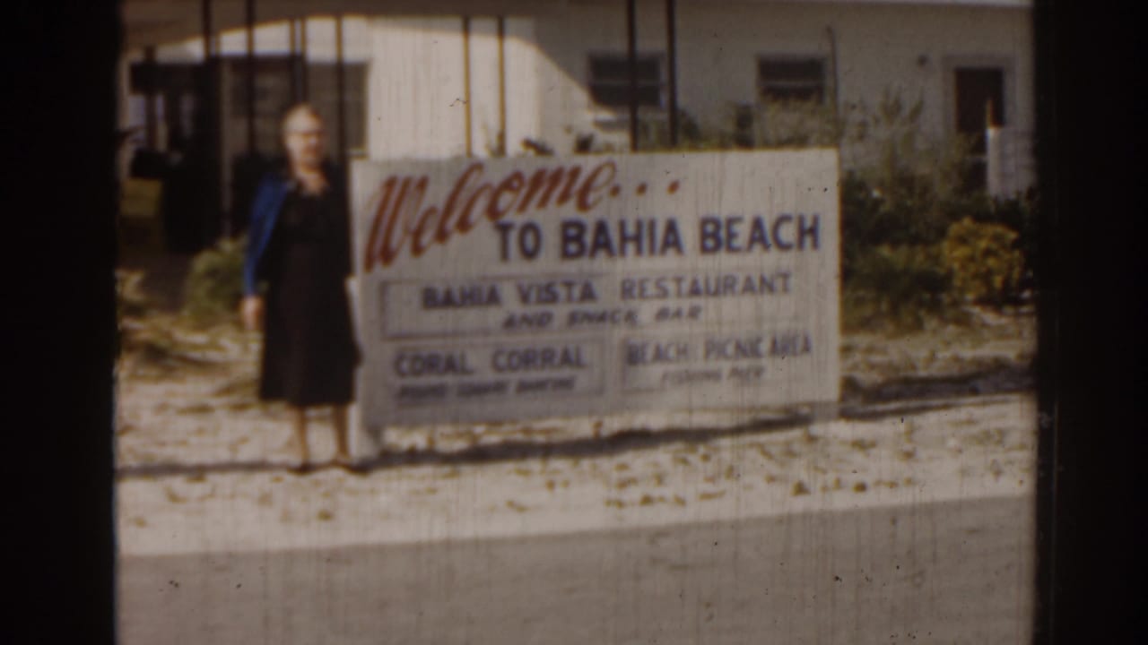 Woman standing beside a large welcome sign for Bahia Beach near sandy ground and low buildings.