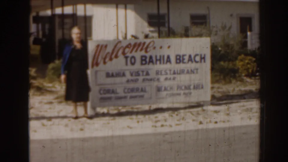 1963 Florida home-movie frame showing an adult woman beside a readable Bahia Beach welcome sign advertising visitor amenities.
