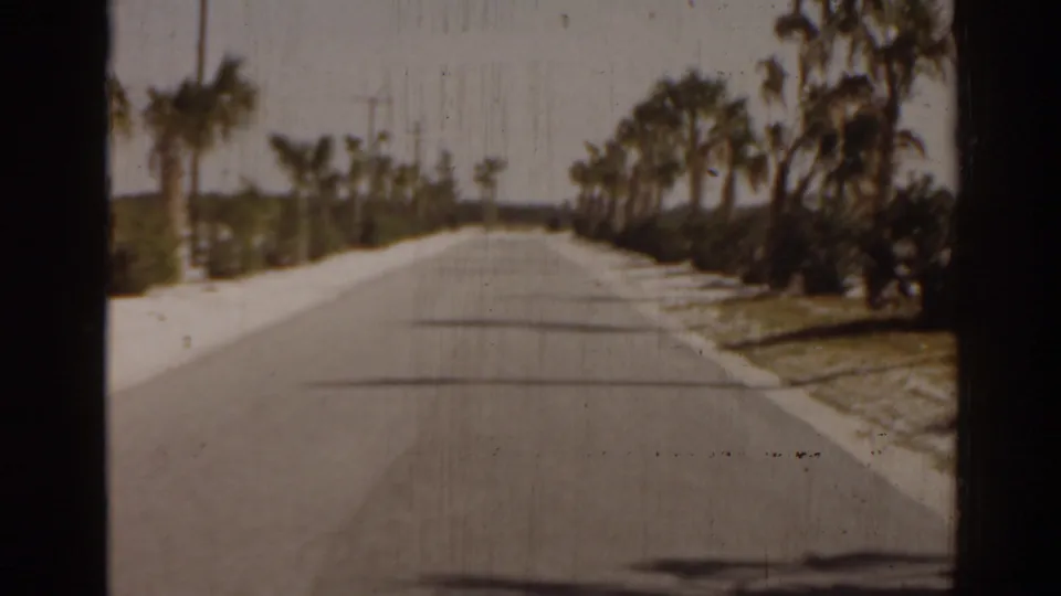 1963 Florida home-movie frame of a straight palm-lined road with sidewalks and tropical planting near a beach destination.