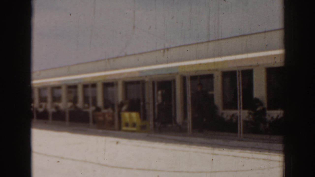 1963 Florida home-movie frame of a building facade, covered walkway, and bright pavement in soft 8mm focus.