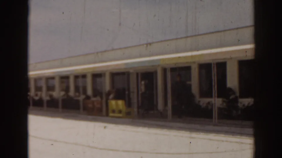 1963 Florida home-movie frame of a building facade, covered walkway, and bright pavement in soft 8mm focus.