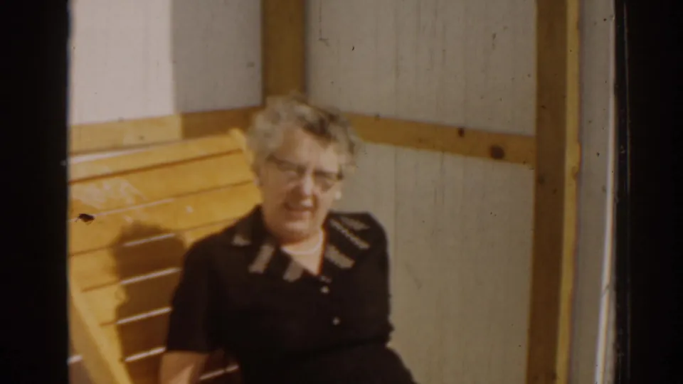 1963 Florida 8mm frame showing an adult woman with eyeglasses seated in a yellow wooden chair beside a paneled wall.
