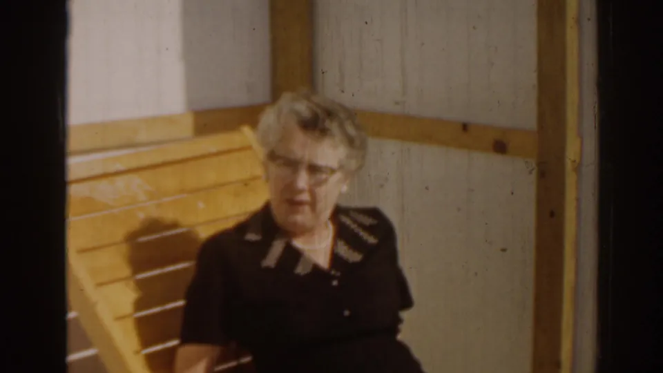 1963 Florida home-movie frame of an adult woman in a black dress seated outdoors against a paneled wall.