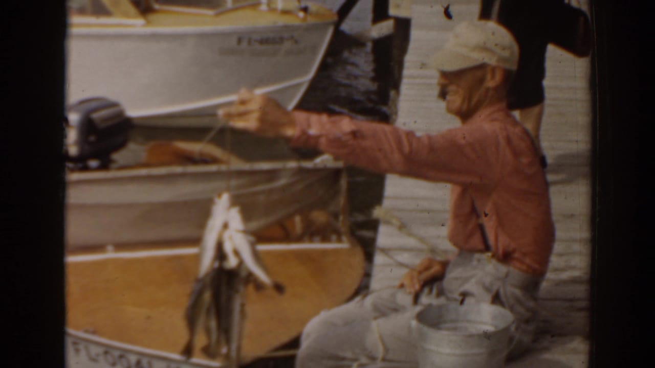 Man in a cap holds up a string of fish beside a small boat on the water.