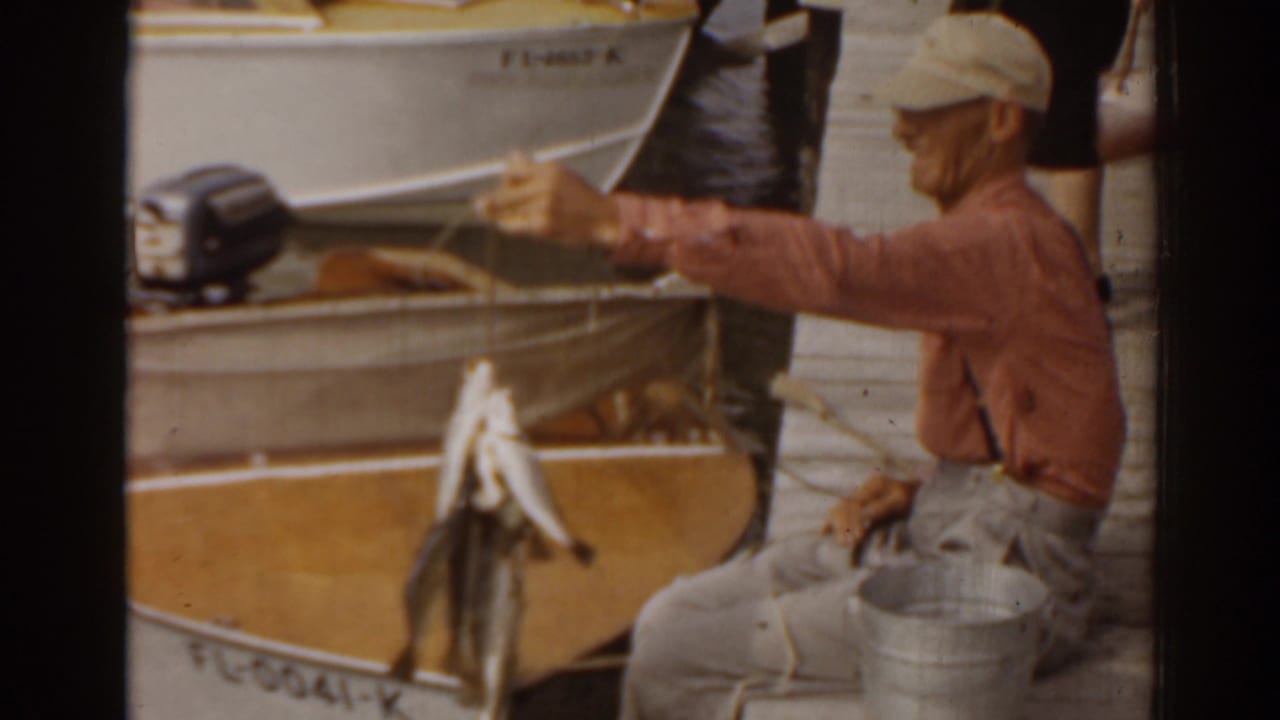 A man in 1963 Florida waterfront footage extends a string of caught fish near a small boat with visible registration markings.