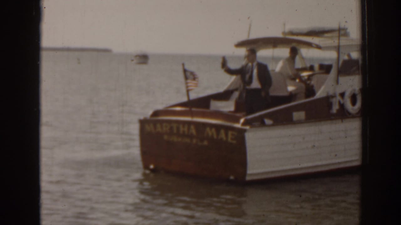 Small motorboat Martha Mae on calm water with a suited man waving and an American flag at the stern.