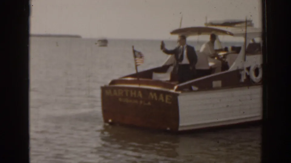 1963 Florida 8mm frame of the Martha Mae motorboat near shore, with a suited passenger waving while another person stands near the helm.