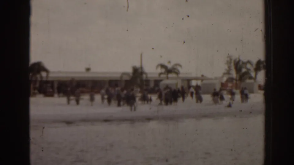 1963 Florida home-movie frame showing a distant public beach or waterfront scene with groups of people near palms and low buildings.