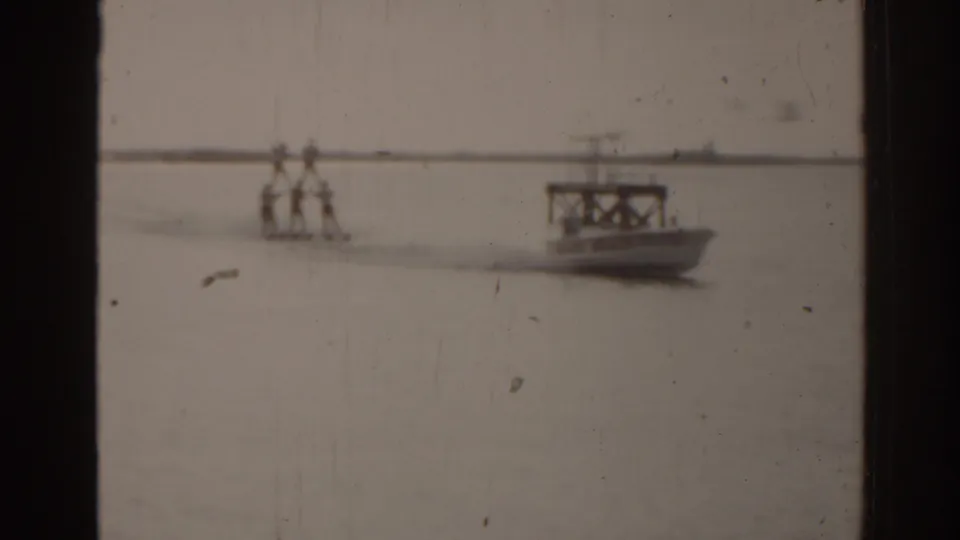 1963 Florida watersports frame with distant water skiers, a small boat, calm water, and soft home-movie texture.
