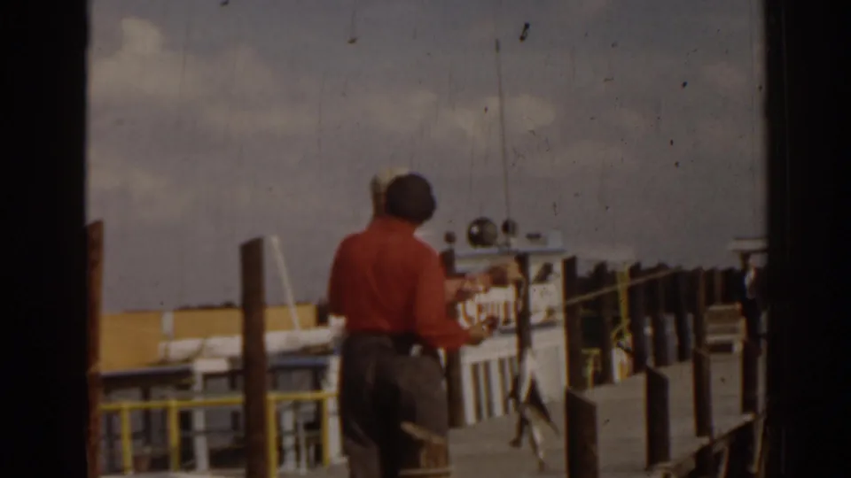 1963 Florida waterfront frame with a man in a red shirt standing on a dock near posts, railings, and water.
