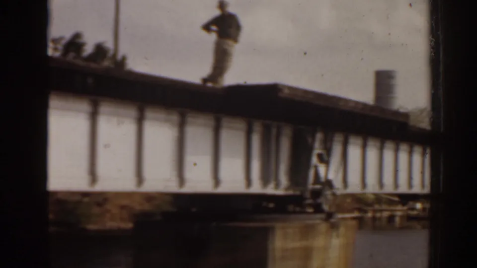 1963 Florida waterfront frame with a person standing on an elevated bridge or pier over water, seen in soft 8mm focus.