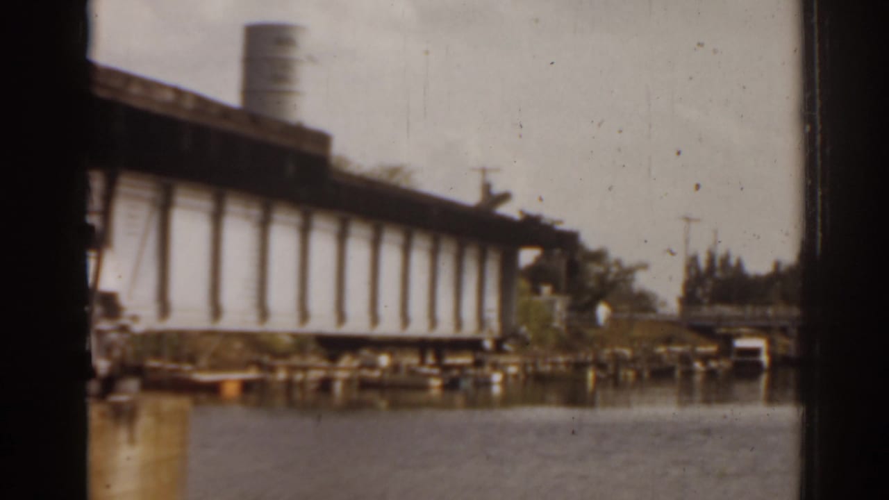 1963 Florida home-movie frame of the drawbridge raised over the river, with a distant shoreline, trees, and small structures visible.