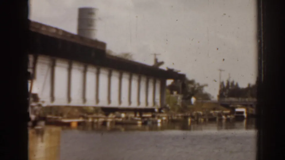 1963 Florida home-movie frame of the drawbridge raised over the river, with a distant shoreline, trees, and small structures visible.