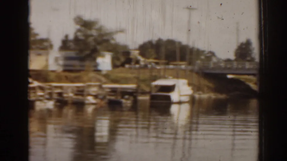 1963 Florida 8mm frame showing a small boat near the bank, shoreline homes, trees, and part of the bridge approach across calm water.