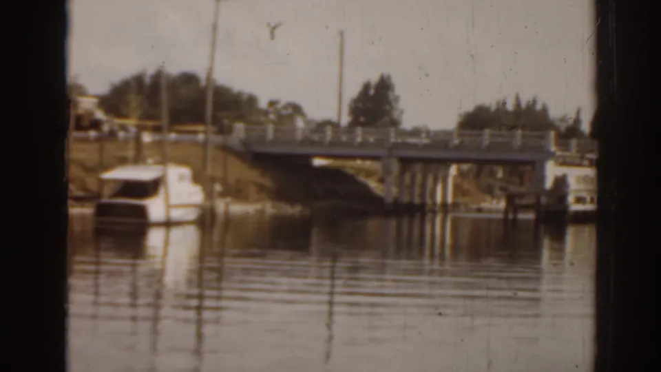 1963 Florida home-movie frame with a low bridge approach, moored small boat, utility poles, trees, and reflections on calm river water.