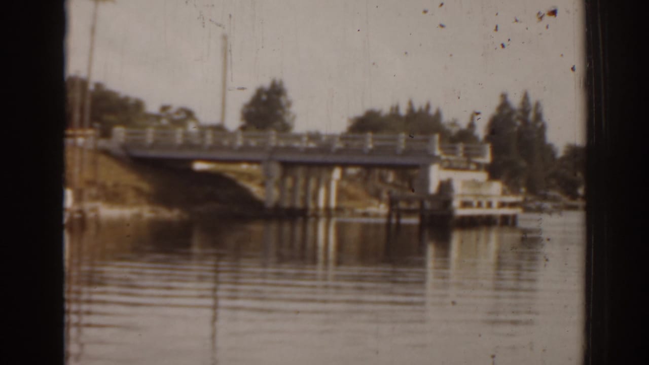 1963 Florida 8mm frame showing a concrete bridge, calm river reflections, and a small white boat along the shore.