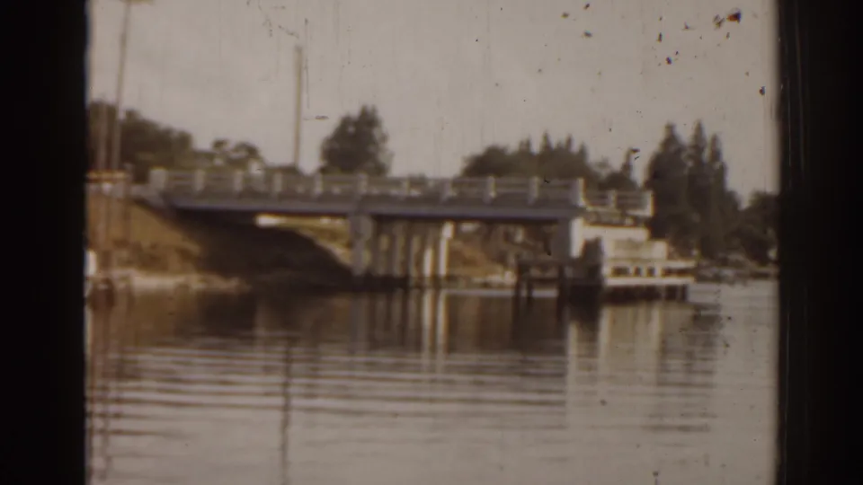 1963 Florida 8mm frame showing a concrete bridge, calm river reflections, and a small white boat along the shore.