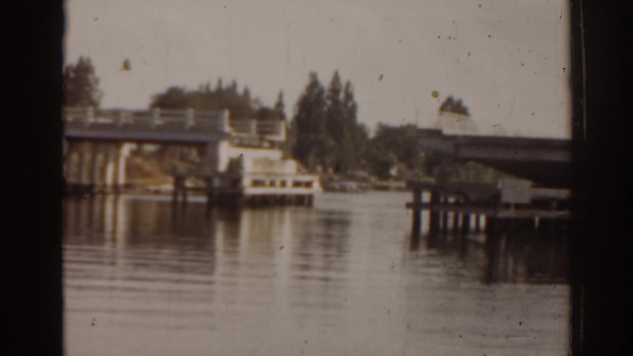 1963 Florida home-movie frame with a bridge, dock pilings, tree-lined shore, and soft reflections on calm water.