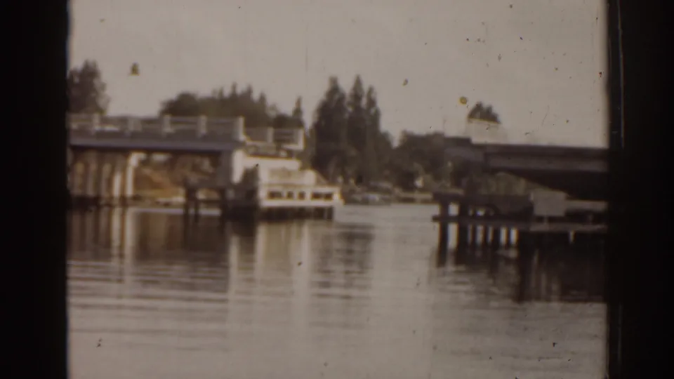 1963 Florida home-movie frame with a bridge, dock pilings, tree-lined shore, and soft reflections on calm water.