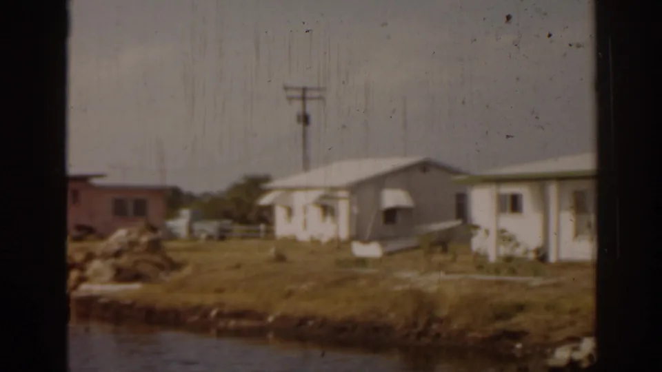 1963 Florida home-movie frame of a modest riverside house, open yard, utility pole, and rocky shoreline.