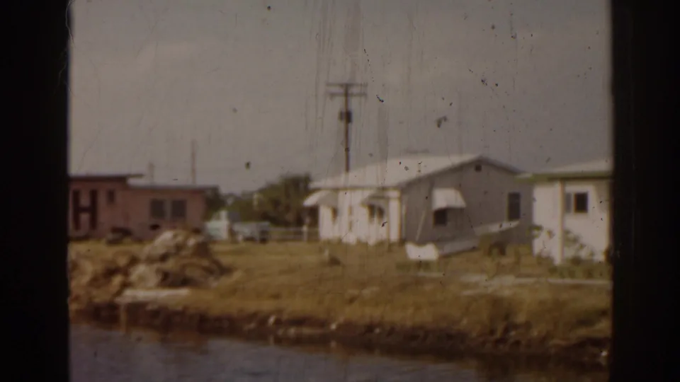 1963 Florida waterfront view with modest buildings, grassy bank, utility pole, and a pink FISH business wall seen through dark frame edges.