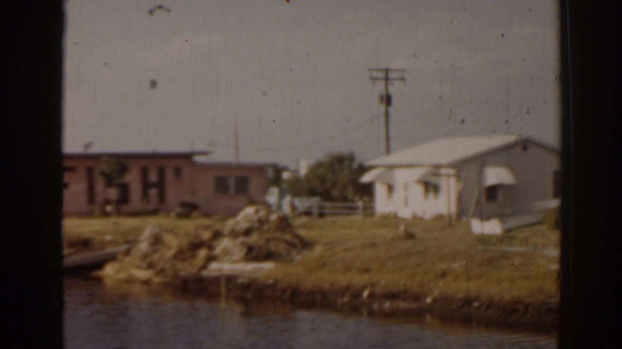 Small waterfront buildings, grassy bank, rocks, and calm water.