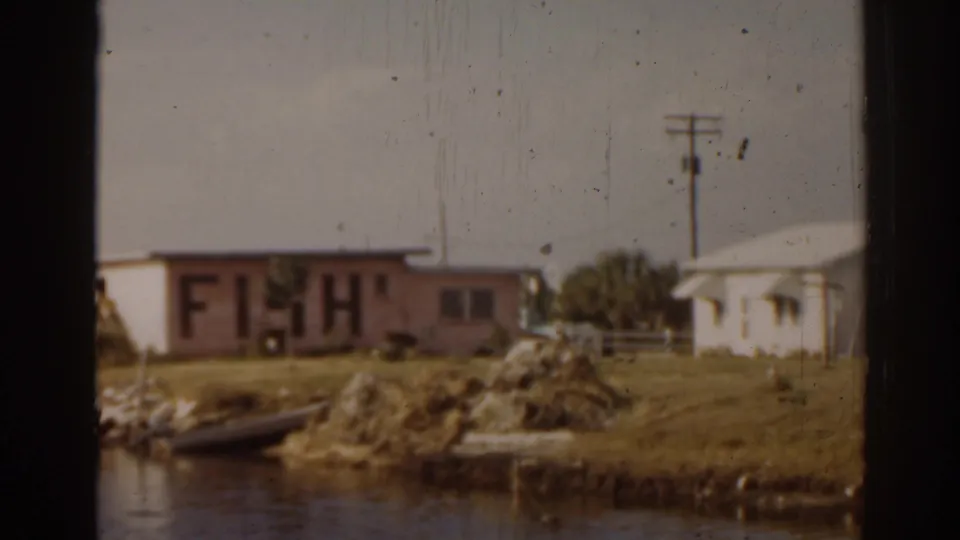 1963 Florida waterfront view showing the FISH-marked building, a small boat on the bank, water in the foreground, and a nearby house.