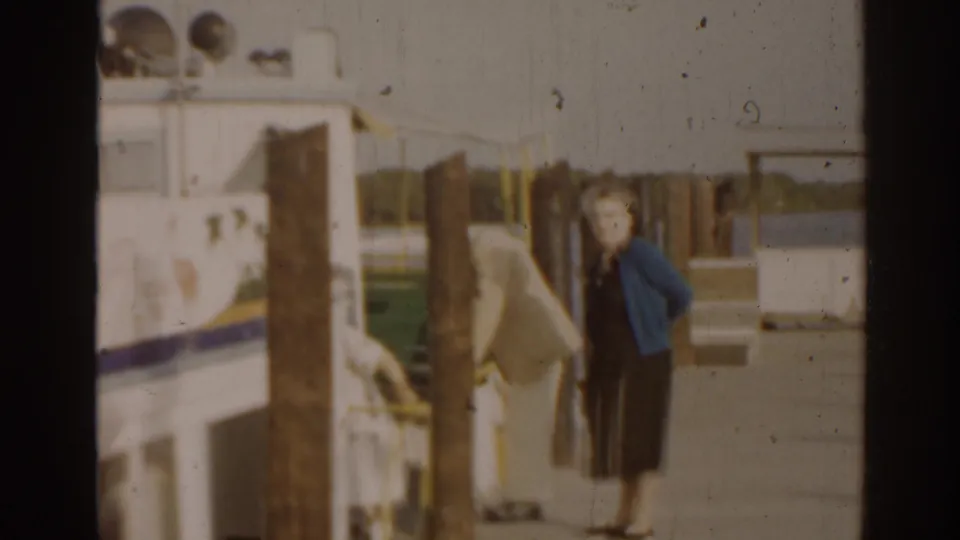 People stand on a dock near a larger vessel, including a woman in a blue cardigan and dark dress in the foreground.