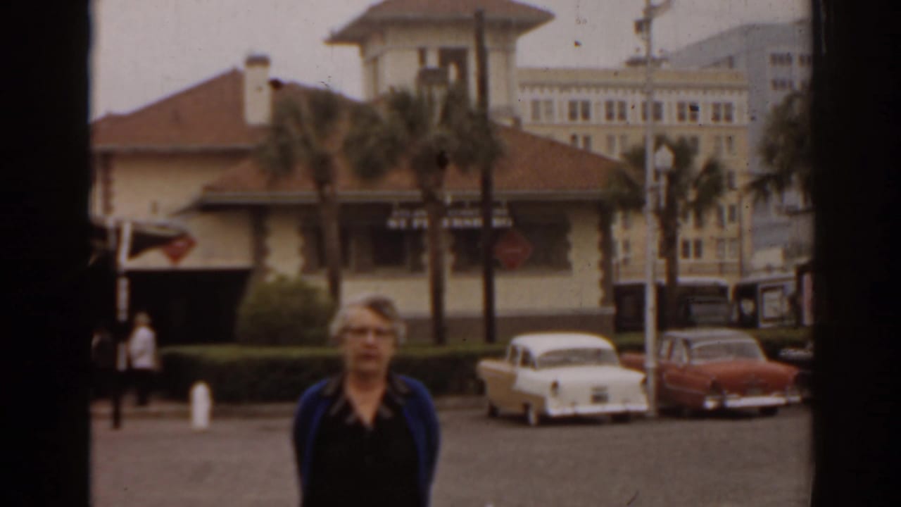Woman in a blue cardigan stands in a Florida parking lot in 1963, with classic cars, palm trees, and a low building behind her.