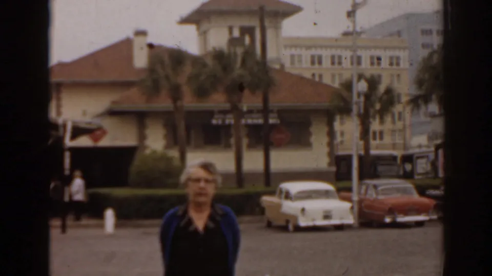 Woman in a blue cardigan stands in a Florida parking lot in 1963, with classic cars, palm trees, and a low building behind her.