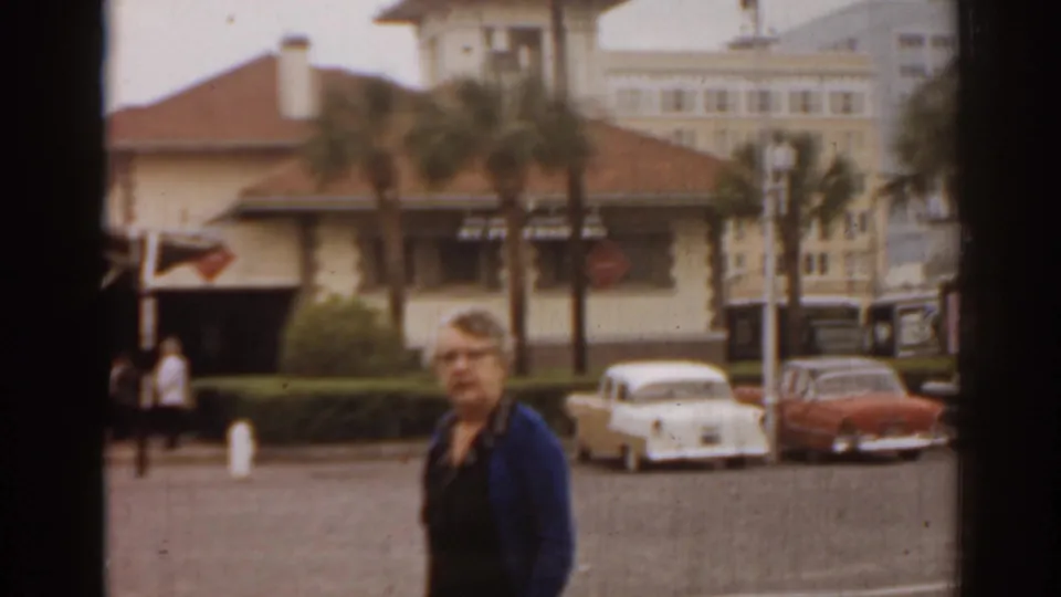 1963 Florida home-movie frame showing a woman beside classic parked cars, palm trees, and a red-roofed street-side building.