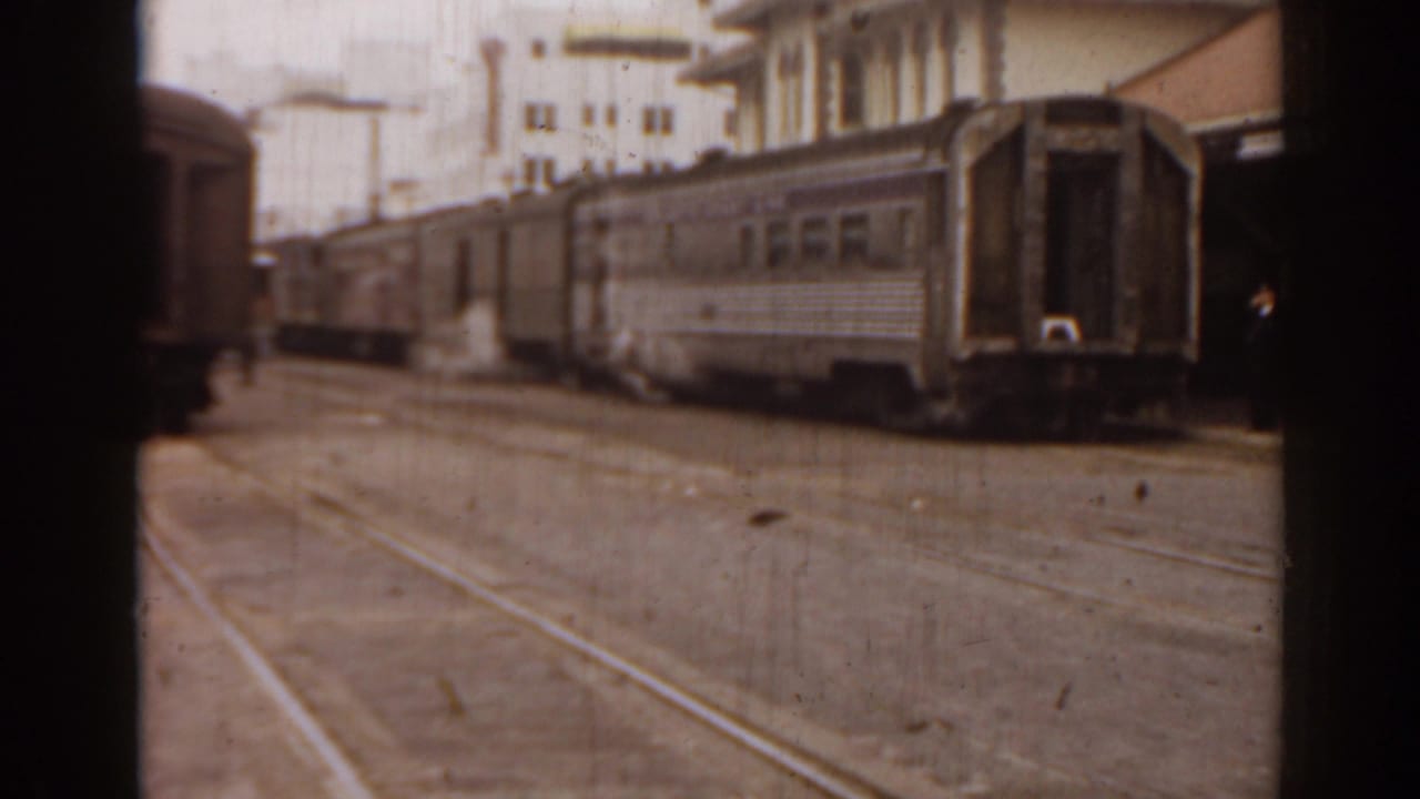Rail cars and paved track area near large buildings, partly framed by dark film edges.