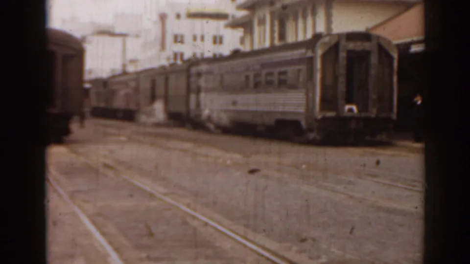 Passenger rail cars stand or pass near a paved rail area and urban buildings in this 1963 Florida home-movie frame.