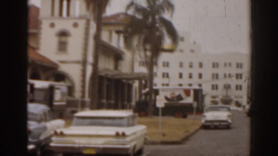 1963 Florida street view with a white sedan, palm trees, and a Spanish-style building with a tower near downtown structures.