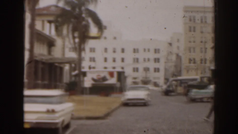 Florida 1963 downtown street scene with moving and parked cars, palm trees, a towered building, and taller buildings in the background.