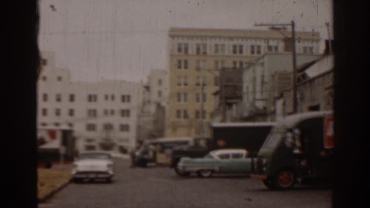 Urban street with older cars, utility poles, buildings, and a delivery truck at the right edge.