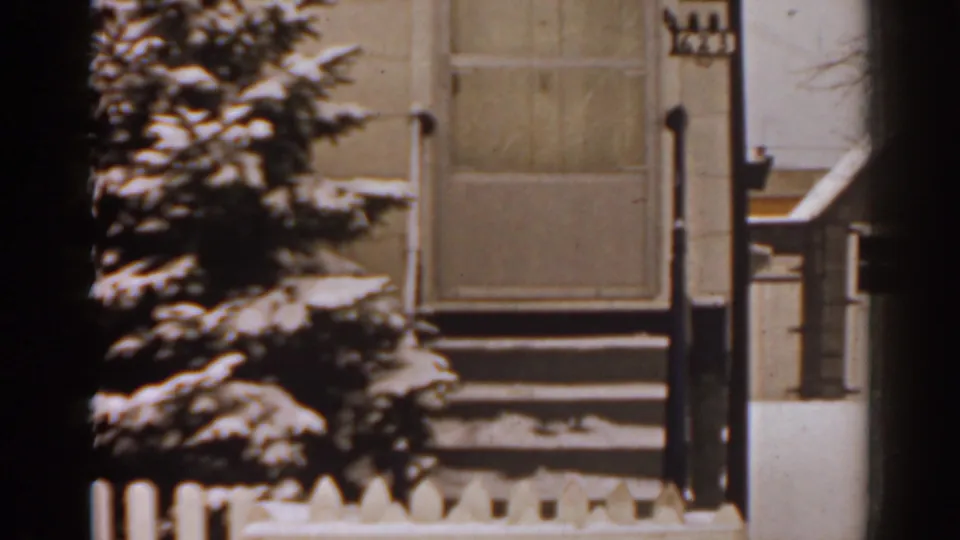 1963 Florida home-movie frame of a snowy house entrance with evergreen branches, porch steps, a door, and white picket fence.