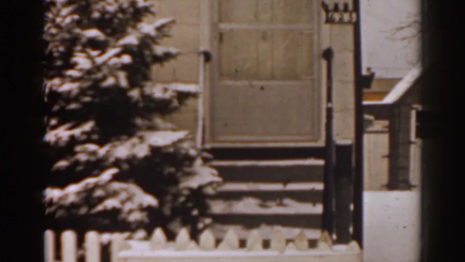 1963 Florida home-movie frame showing snow on porch steps, a picket fence, and an evergreen near a house exterior.