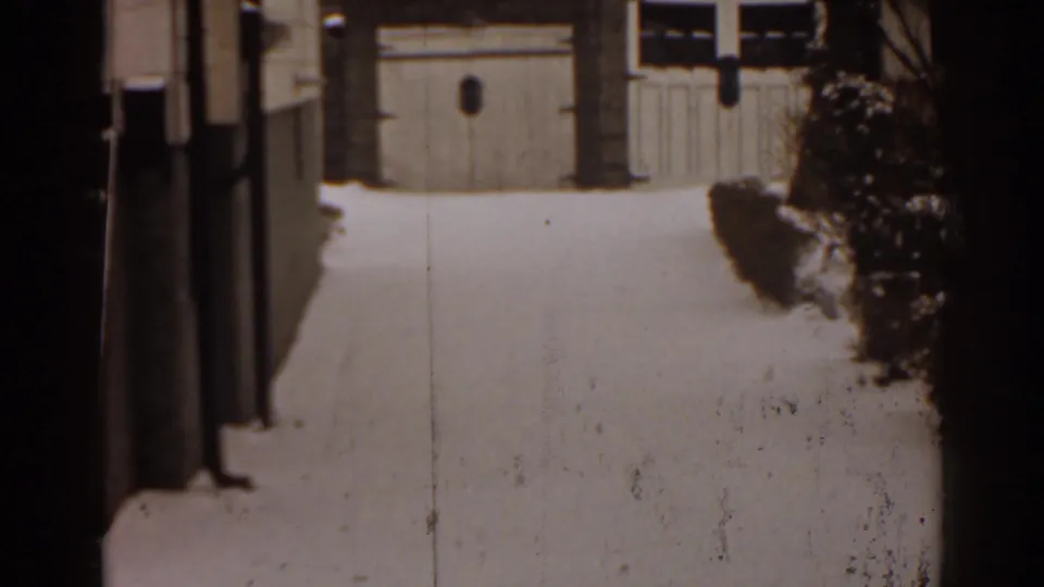 1963 Florida frame showing a snow-blanketed residential yard with garage doors, posts, and shrubs in view.