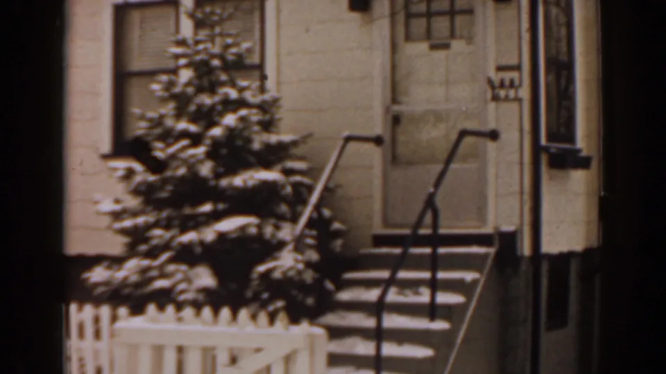 1963 Florida home-movie frame showing a snow-dusted suburban front stoop, small evergreen, white fence, and black metal railings.